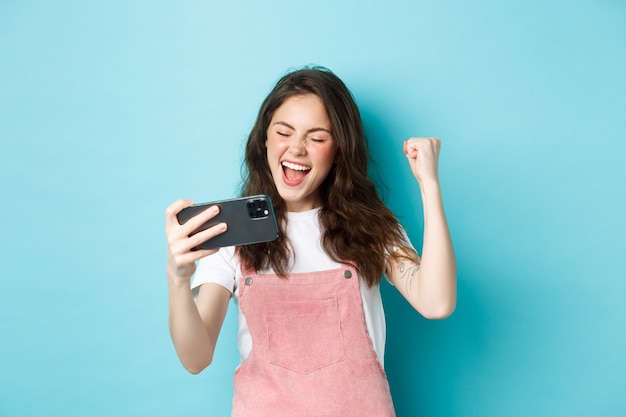 Cheerful cute girl winning in online video game on smartphone, making fist pump and shouting yes with joy, standing over blue background and triumphing.