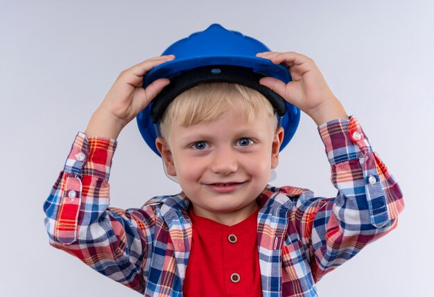 A cheerful cute little boy with blonde hair wearing checked shirt holding his blue helmet while looking on a white wall