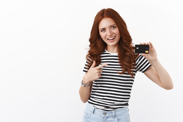 Cheerful cute redhead girl opened deposit gather money for summer holiday, pointing at black credit card and smiling joyfully, paying online, using cashback to pay for coffee