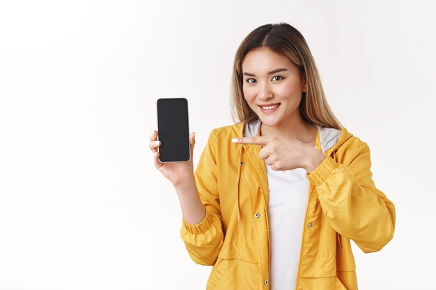 Веселая милая нежная азиатская блондинка в желтой стильной куртке держит показ смартфона, указывая указательным пальцем на экране телефона, широко улыбаясь, рекомендую крутое приложение, стоящее на белой стене
