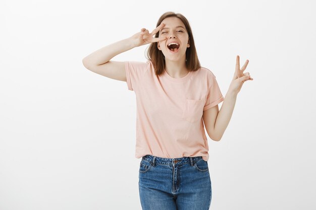 Cheerful cute woman showing peace gestures, standing kawaii over white wall