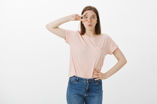 Cheerful cute woman showing peace gestures, standing kawaii over white wall