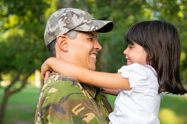 Cheerful dad in camouflage uniform holding little daughter in arms, hugging girl outdoors after returning from military mission trip. Closeup shot. Family reunion or returning home concept