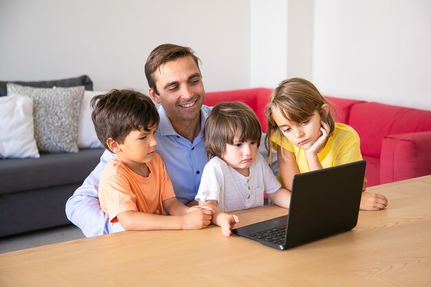 Cheerful dad and pensive kids watching movie via laptop together during weekend. Happy father sitting at table with children in living room. Fatherhood, childhood and digital technology concept