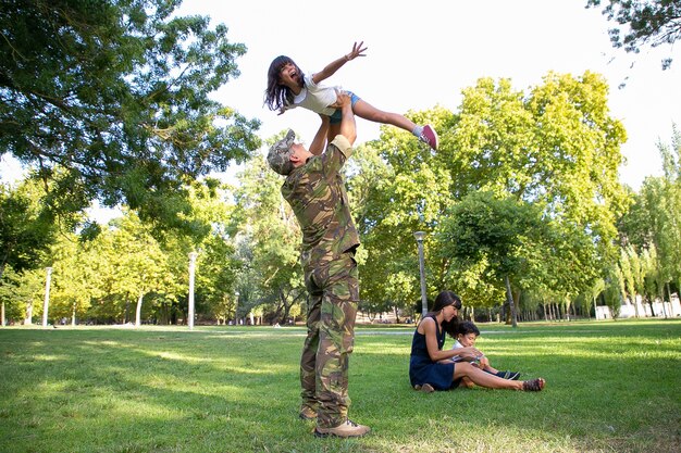 Cheerful dad rising up his daughter and standing on lawn. Happy father playing with exited girl in park. Brunette mom and little son sitting on grass. Family reunion and returning home concept