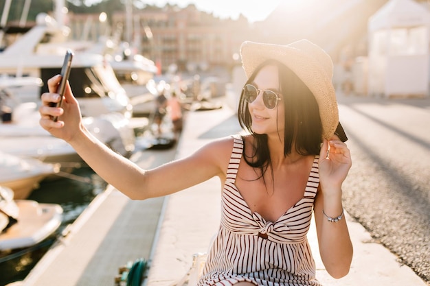 Cheerful dark-haired girl in sunglasses making selfie while resting in river port enjoying sunshine. Outdoor portrait of smiling young woman in hat and dress taking picture of herself near yachts.