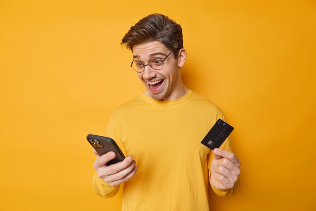 Cheerful dark haired man holds smartphone and credit card involved in online shopping purchases goods or purchases in internet store wears spectacles casual jumper isolated over yellow wall.