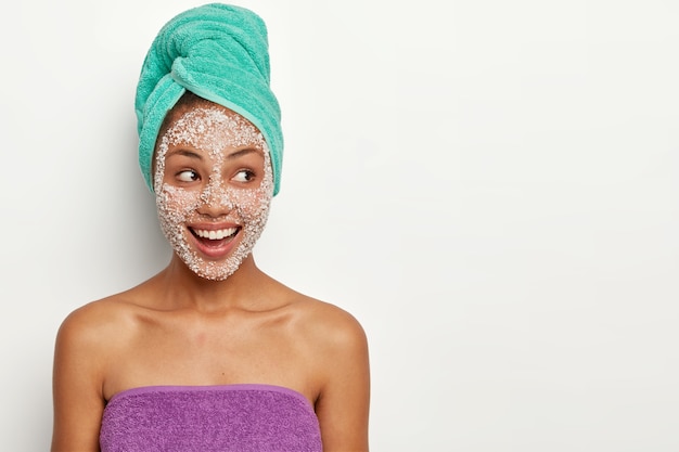 Free photo cheerful dark skinned female model wears towel on head and body, applies sea salt mask for peeling from pores, looks aside with satisfaction