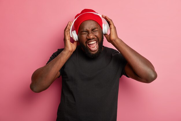 Cheerful dark skinned hipster man uses headphones for noise cancellation, listens rock music, sings song aloud, wears red hat and black t shirt.