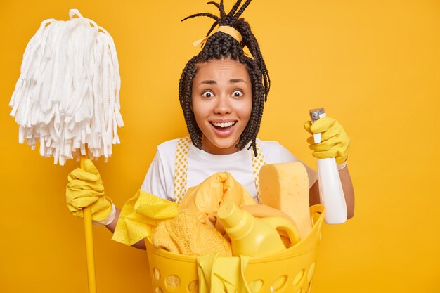 Cheerful dark skinned housewife holds chemical detergent and mop looks gladfully