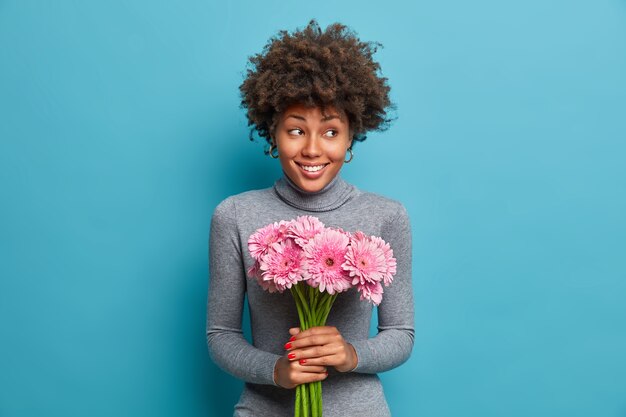 Cheerful dark skinned lady holds bouquet of pink gerbera daisy, looks with delight and happiness aside, wears grey turtleneck