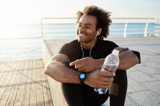 Cheerful dark-skinned muscular athlete in black sport clothing sitting on pier after sport activities wearing white earphones.
