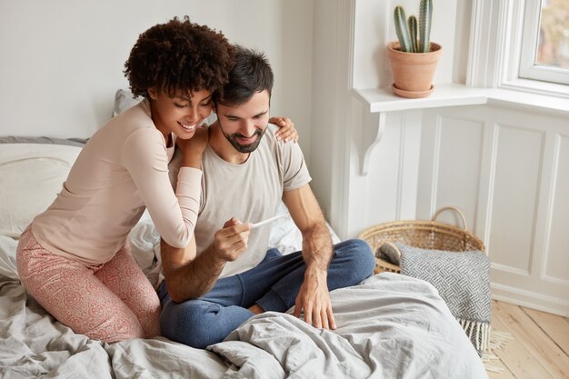 Cheerful dark skinned woman embraces her husband, shows positive result on test, rejoice they will become parents soon,