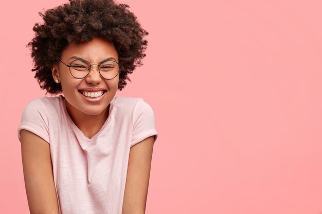 Cheerful dark skinned woman giggles, shrugs shoulders, squints face, dressed casually, models in against pink wall