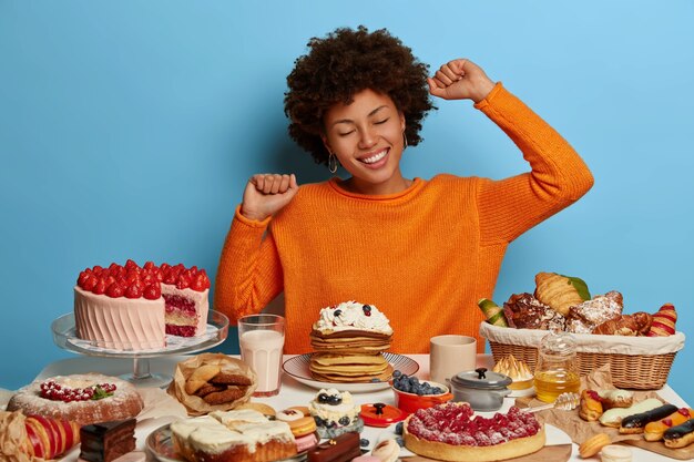 Cheerful dark skinned woman stretches hands, dressed casually, sits at table with many tasty cakes, desserts and pies, being in good mood as eats delicious food