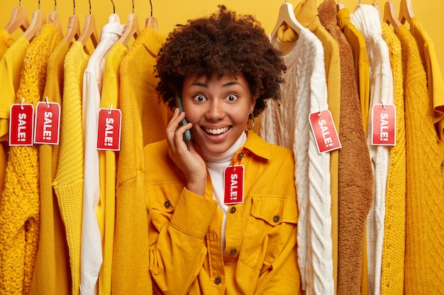 Cheerful dark skinned woman with charming smile, stands between clothes with red tags inscriped sale,  look gladfully at camera