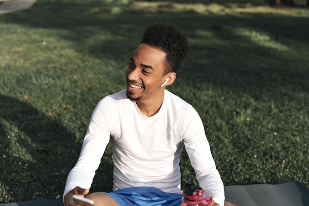 Free photo cheerful darkskinned bearded guy in white longsleeved tshirt and blue shorts smiles listens to music in headphones holds phone and water bottle