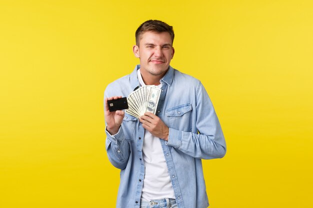 Cheerful and delighted happy blond guy looking excited, smiling satisfied as receive extra cash payment, holding credit card with money, standing yellow background upbeat.