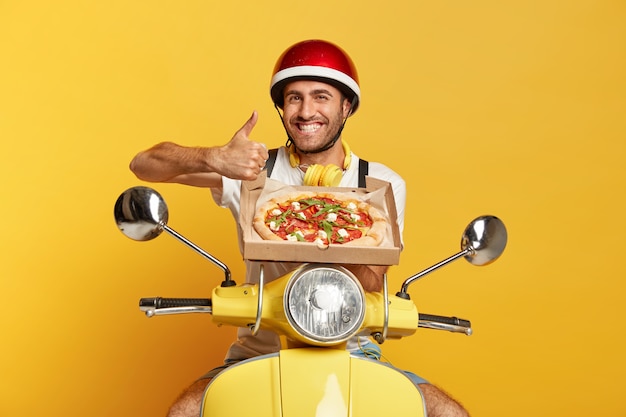 Cheerful deliveryman with helmet driving yellow scooter while holding pizza box