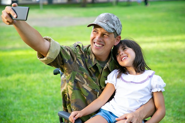 Cheerful disabled military dad and his little daughter taking selfie together in park. Girl sitting on dads lap. Veteran of war or disability concept