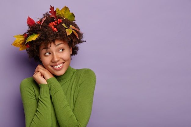 無料写真 陽気な夢のようなアフリカ系アメリカ人の女の子は頭を傾け、心地よく微笑んで、脇を見て、屋内で肖像画を作り、黄色の葉、髪にナナカマドの果実を持っています