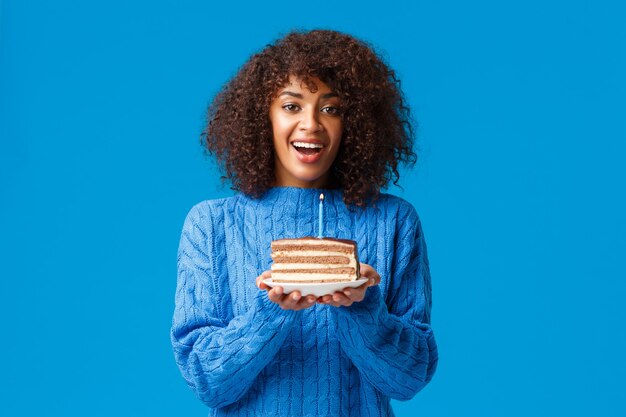 밝고 꿈꾸는 귀여운 아프리카 계 미국인 b-day 소녀, 촛불 케이크를 들고 불고 웃고, 생일 파티, 스웨터 파란색 벽에 서.