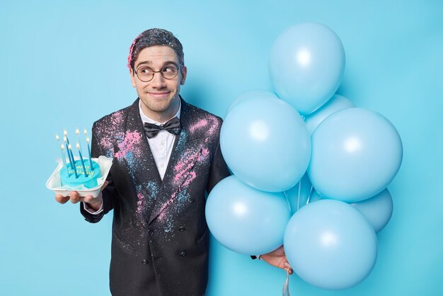 Cheerful dreamy young man dressed in formal clothes looks away holds sweet cake with burnig candles and bunch of inflated balloons ready for birthday celebration isolated over blue background