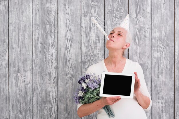 デジタルタブレットと紫の花の花束を手で押しパーティーホーンを吹いている陽気な年上の女性