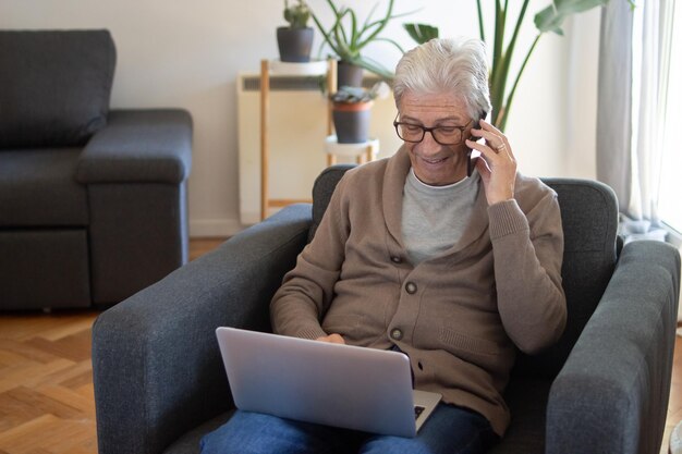 Libero professionista anziano allegro che lavora a distanza. uomo sorridente dai capelli grigi in cardigan che usa il laptop mentre chiama per telefono. concetto di affari online