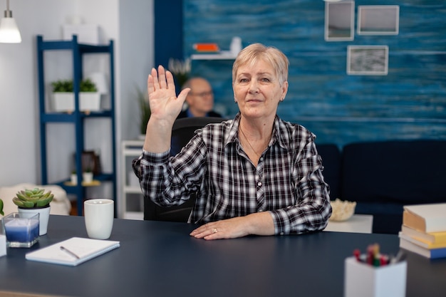 Cheerful elderly pensior waving at camera and husband is reading a book sitting on sofa. Happy senior woman with gray hair saying hello at camera during video call in home living room.