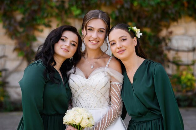 Cheerful elegant bride young woman posing with two bridesmaids in dark green dress standing outdoors Attractive woman in wedding dress with long sleeves Celebration of wedding Friends of bride