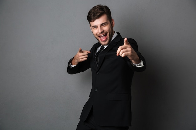 Cheerful emotiojnal young man in suit poiting with two finger on you, 