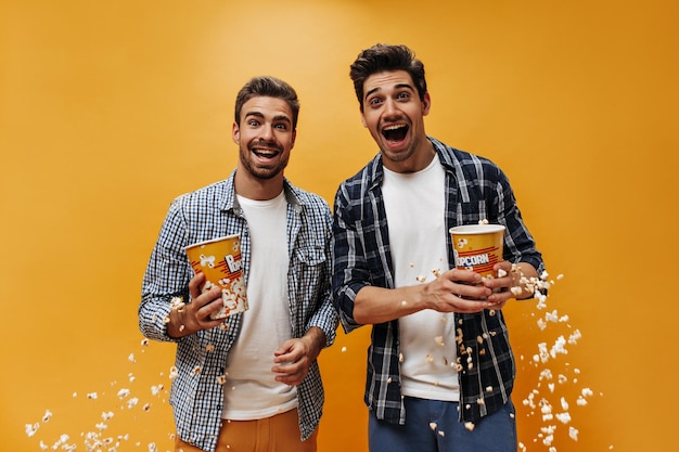 Веселые эмоциональные счастливые мужчины в укороченных рубашках и белых футболках искренне улыбаются, держат попкорн и позируют на оранжевом фоне