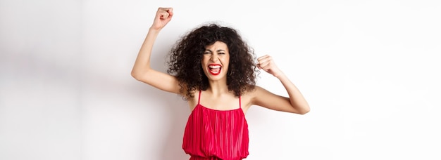 Free photo cheerful emotive woman with curly hair red dress raising hands up and chanting rooting for team shou