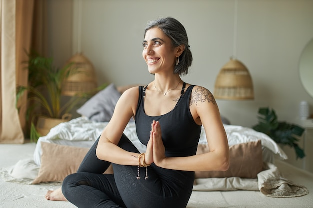 Cheerful energetic young female sitting in spinal twist on floor at home, doing ardha matsyendrasana during yoga practice