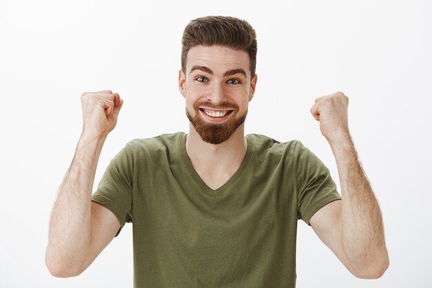 Cheerful and energized active cute male fan with beard in t-shirt raising clenched fists in victory and triumph celebrating win of first prize smiling from excitement and delighted over white wall