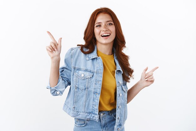 Cheerful enthusiastic modern young redhead female in denim pointing sidweays showing left and right copy space advertising offers smiling and laughing carefree recommend good quality services