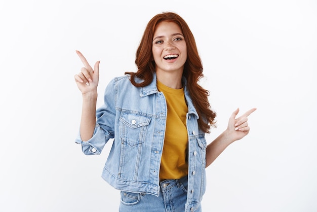 Cheerful enthusiastic modern young redhead female in denim pointing sidweays showing left and right copy space advertising offers smiling and laughing carefree recommend good quality services