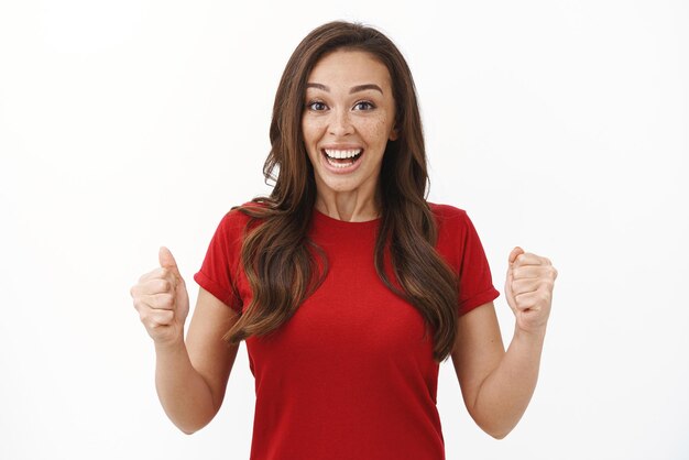 友人の拳ポンプの笑顔を応援している赤いTシャツの陽気な熱狂的な若いブルネットの女性は達成された成功を達成します