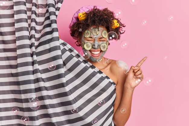 Жизнерадостная этническая женщина наносит маску красоты на лицо, показывает, что, если не принимать во внимание позы в ванной, собирается принять душ, хихикает, положительно имеет чистое тело, здоровую ухоженную кожу.