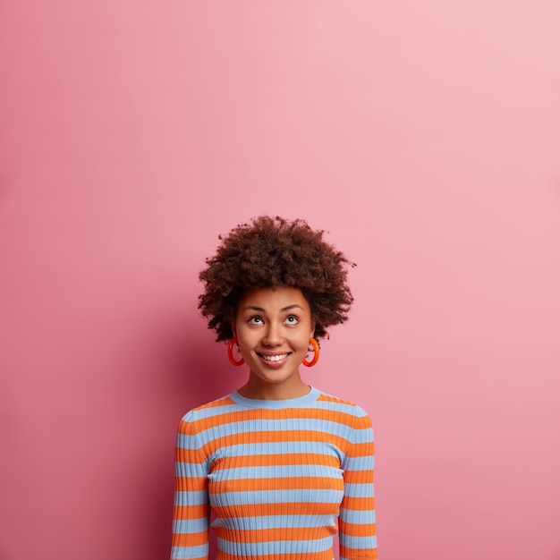 Cheerful ethnic girl with curly hair, concentrated above, being in good mood, looks curiously upwards, dressed in casual striped jumper, isolated on pink wall, empty space