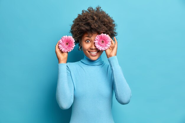 Cheerful ethnic woman holds gerberas in front of eyes smiles positively enjoys pleasant aroma of flowers