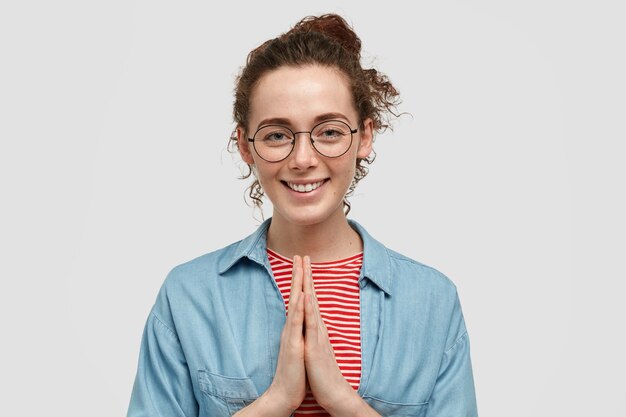 そばかすが顔に付いている陽気なヨーロッパの女性は、祈りのジェスチャーで手を保ち、何か良いものを信じて、白い壁に隔離された優しい笑顔を持っています。