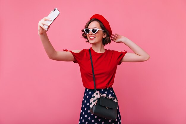 Cheerful european girl with cute tattooes making selfie. Wonderful french woman in beret and sunglasses taking picture of herself.