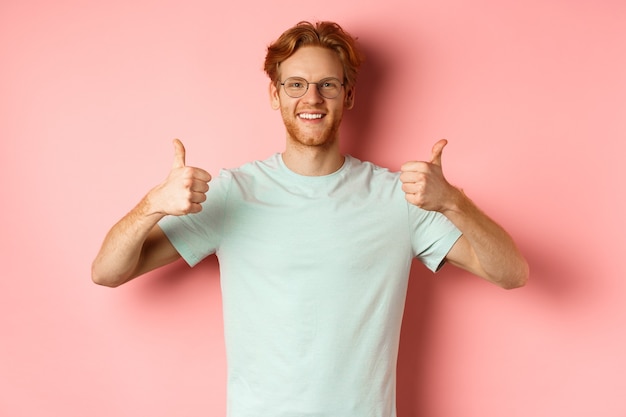 Cheerful european man with red hair and beard wearing glasses showing thumbsup and smiling in approv...