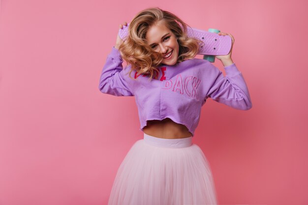 Cheerful european woman holding her skateboard and funny laughing. Amazing blonde girl having fun on pastel.