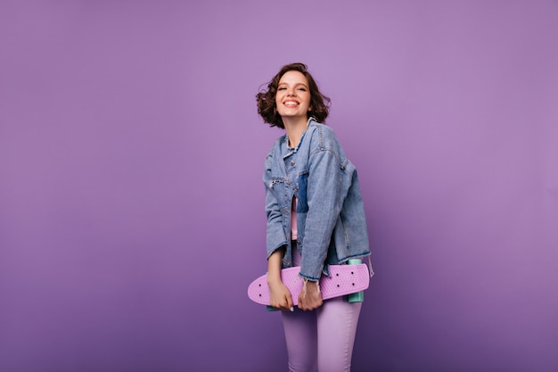Cheerful european woman in purple pants posing with skateboard. Indoor shot of appealing smiling girl with dark wavy hair.