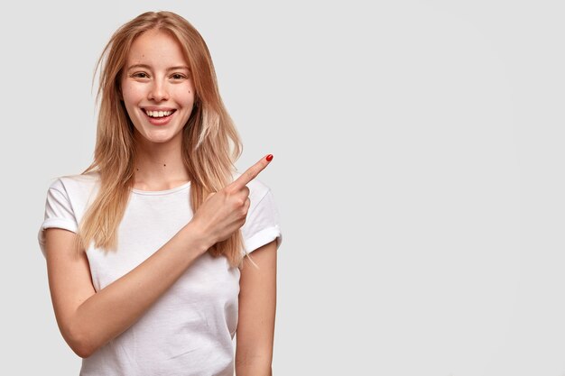 Cheerful European woman with broad smile, appealing look, points aside, dressed in casual white t shirt, shows something pleasant, advertises new item in shop, copy space for your text or promotion