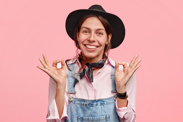 Cheerful European woman with positive smile, makes okay gesture with both hands, demonstrates her agreement and rejoices her success, poses against pink wall