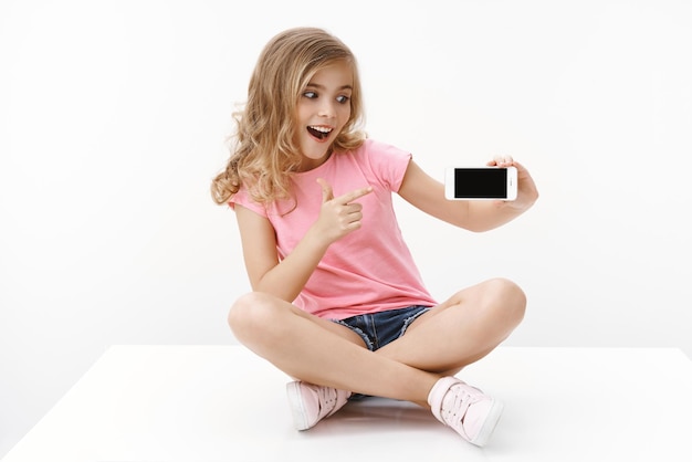 Cheerful excited cute blond little girl sitting on floor with crossed legs hold smartphone pointing mobile phone display and looking thrilled cellphone surprised showing favorite internet page